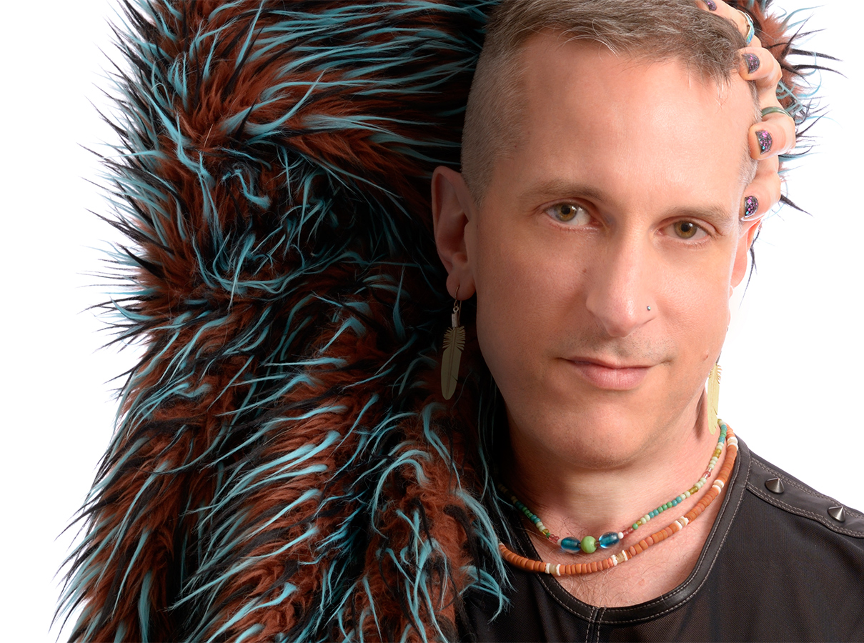 DJ Princess Petite, a white man with close-cut hair, wearing a black t-shirt with spikes. His right arm in raised behind his head so that you see his speckled nail polish. He's wearing a fake fur in black, rust, and neon blue over that arm.